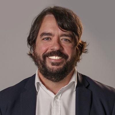 Headshot of Trent Burke, He is wearing a white shirt a black jacket and standing behind a grey wall. 