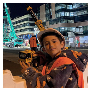 A boy in a hard hat and fluorescent orange vest holds a Lego crane in front of the New Footscray Hospital construction site