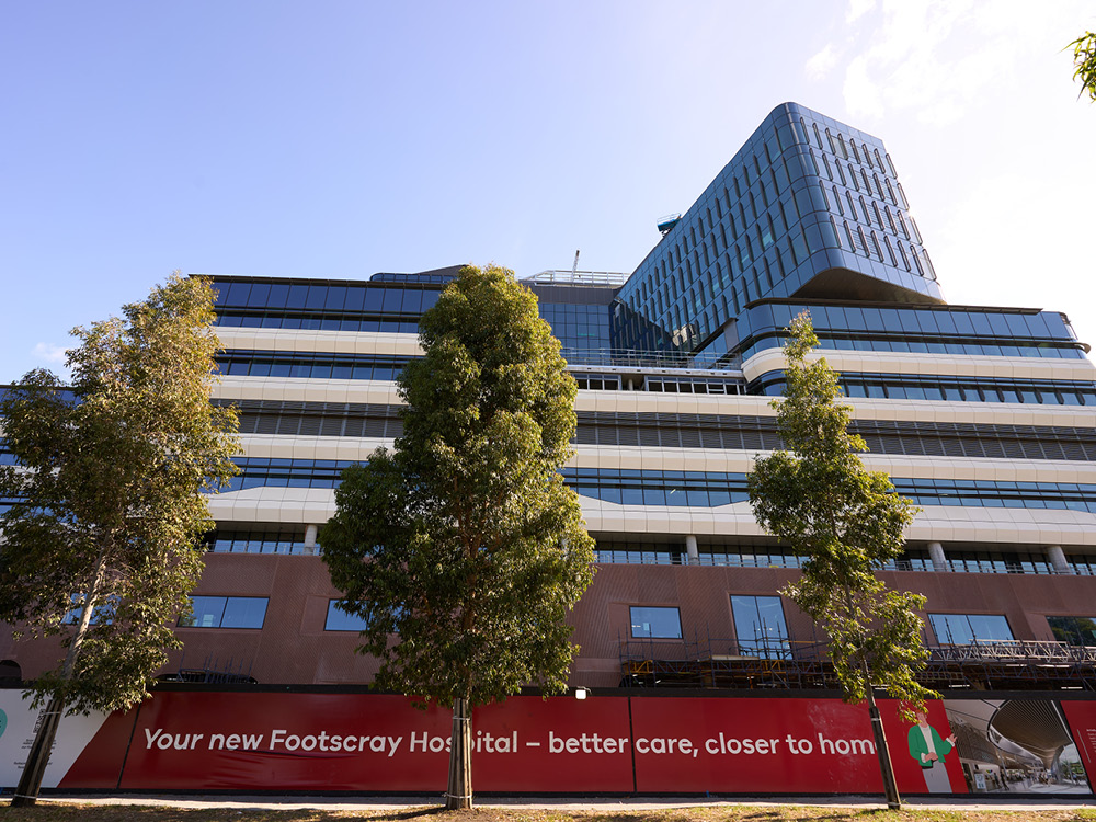 A view from the street looking up at the facade of New Footscray Hospital. In the foreground, three trees stand in front of signage that reads "Your new Footscray Hospital - better care, closer to home"