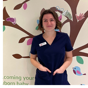 A woman smiles infront of a colourful tree wall decal with a sign that reads 'Birth Tree: Welcoming your newborn baby'