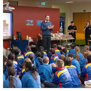 A man stands on stage talking animately to a group of sitting schoolchildren