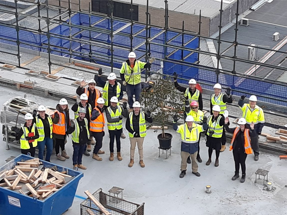 Aerial shot of eighteen people in fluorescent vests and hard hats waving at the camera, while standing on a construction site