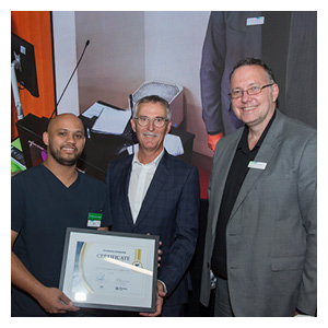 Three people standing, smiling at the camera. The person on the left holds a certificate.