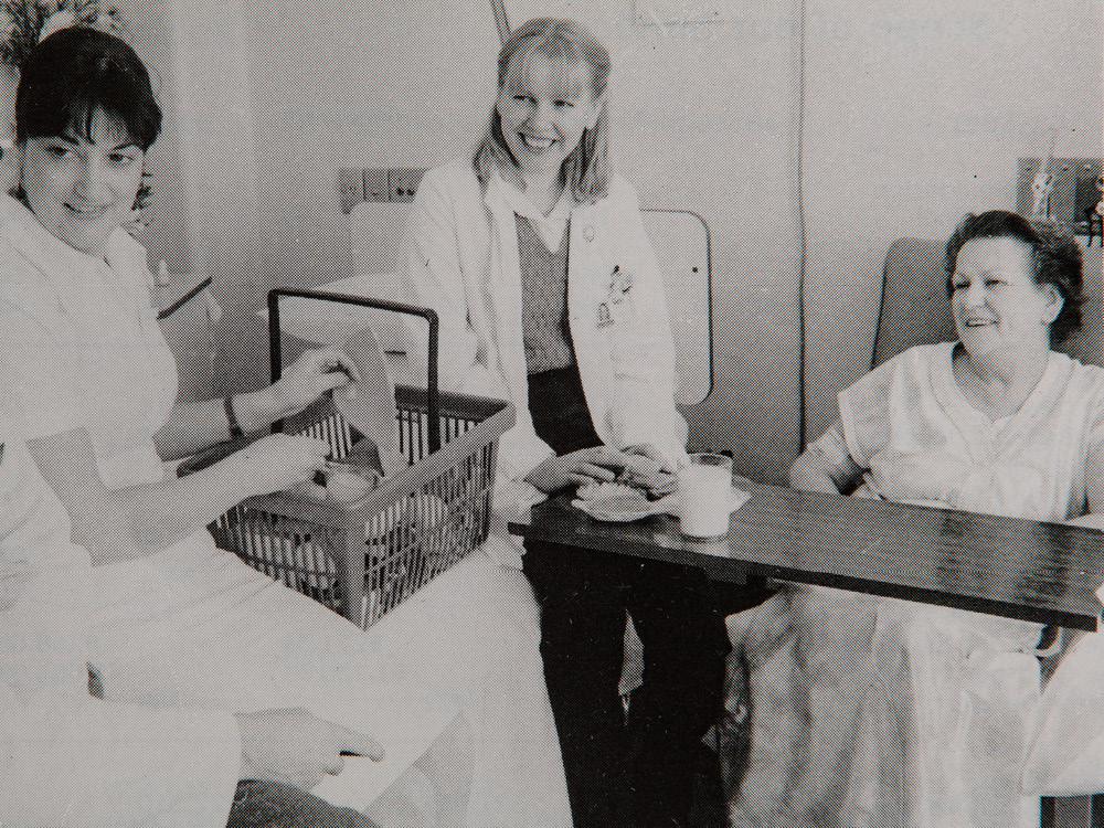 A doctor and a nurse sit with a patient, smiling. The image is in black and white, taken in the 1980s.