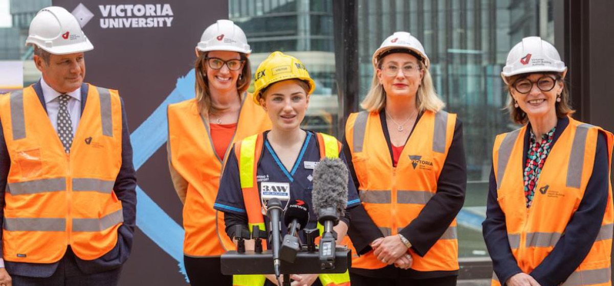 Four people in hard hats and orange fluorescent vests stand behind a nursing student wearing a yellow hard hat and orange vests speaking into a microphone stand