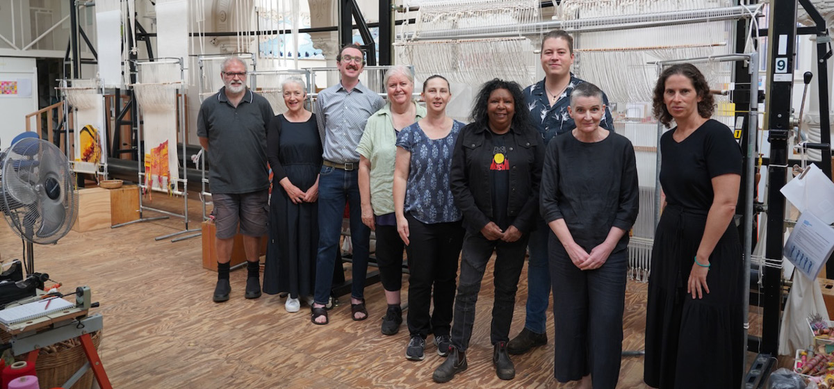 Nine people stand smiling at the camera in a tapestry workshop