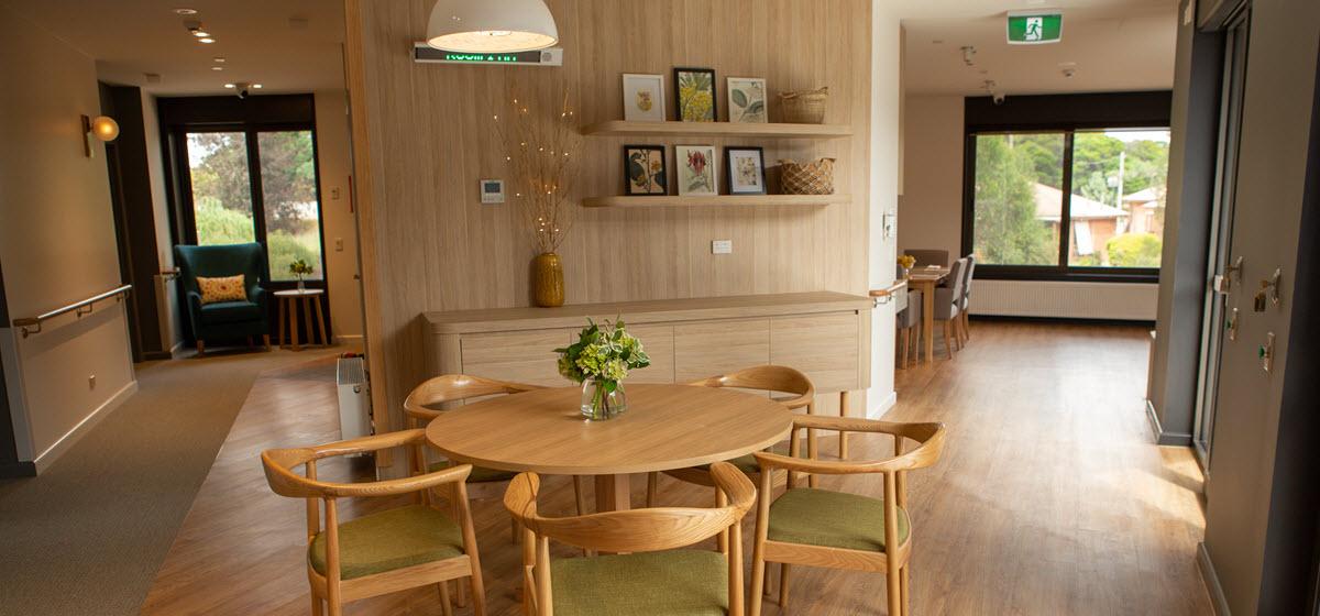 A living room at Creswick Nursing Home, with a wooden table and chairs, shelves with pictures on the wall and soft lighting