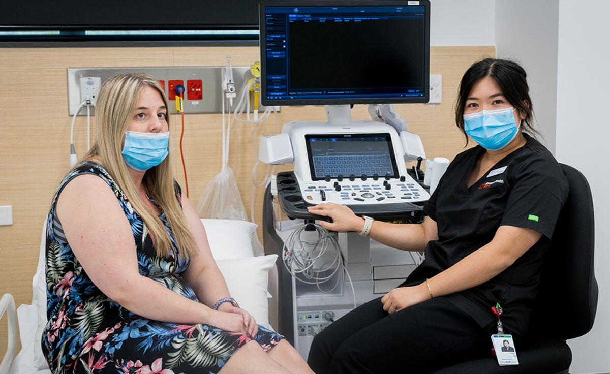 A patient wearing a mask sitting on a bed with a medical professional at the Victorian Heart Hospital