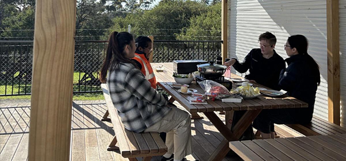 Four people sitting at a timber table on outdoor timber decking, enjoying a meal together