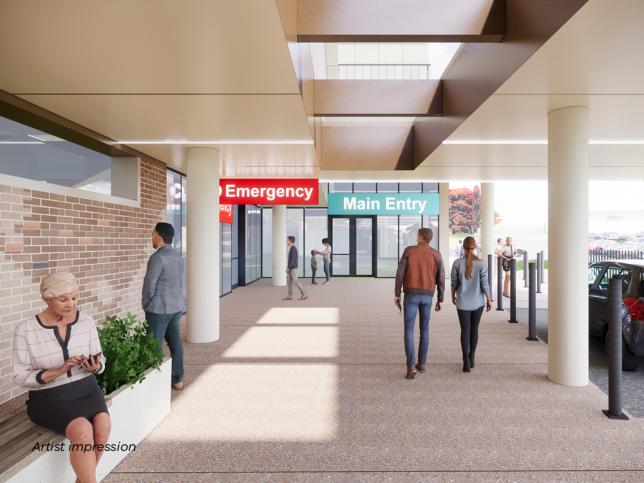 People walking and sitting at the main and emergency entrance of the Wonthaggi Hospital. There are two pillars, a bench and a sign that says 'Emergency'