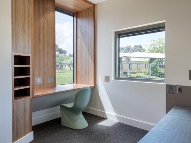 A private room at the Thomas Embling Hospital. There are two windows and a green chair next to a desk.