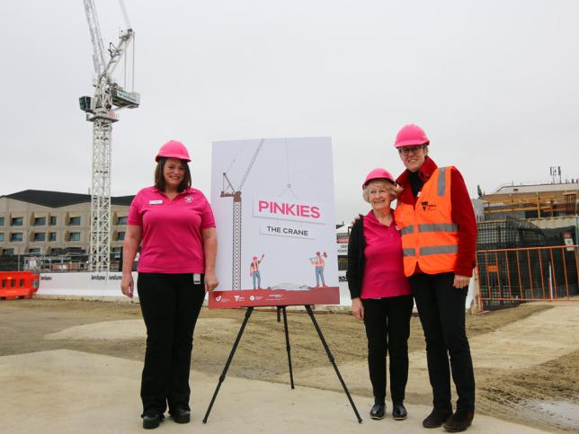 Three women wearing pink hard hats standing in front of a sign that reads 'Pinkies the crane' at the Frankston Hospital redevelopment site.