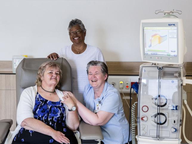 One woman sitting on and two women standing beside a renal dialysis chair and machine