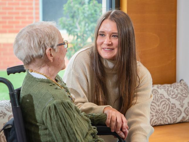 A young women sits beside an older woman in a chair, holding her hand and smiling