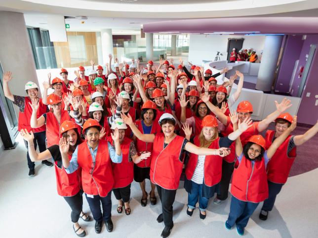 A large group of women in red vests and hardhats smiling and waving their hands in the air