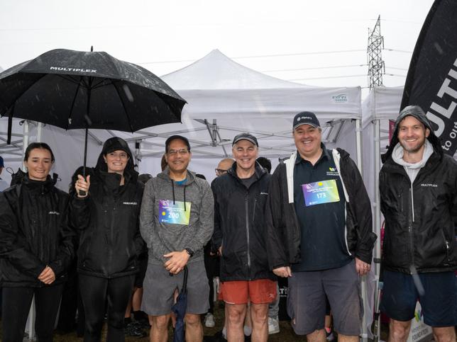 Six people standing and smiling at the camera. Some are wearing black rain jackets with the Multiplex logo on them, and one person is holding an umbrella