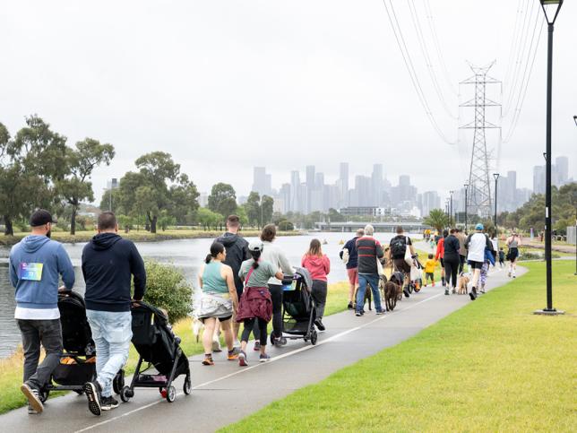 A large group of people walking along a path next to a river. Some are pushing prams