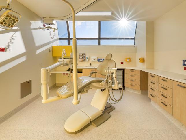 A dental consulting room with white walls and floor, and a large window at the back of the room. A dental chair sits in the middle of the room, with draws and bench tops along the walls