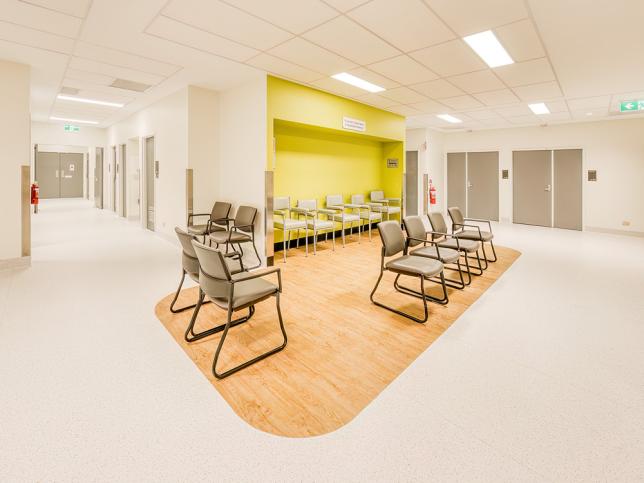 A waiting room with white walls and floors. The seating area has a yellow wall, timber flooring and brown chairs