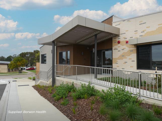 The Geelong EPC entrance from the perspective of somebody standing on the footpath. The accessible concrete ramp and metal rail leads to the glass doors, which are surrounded by brown and white facade