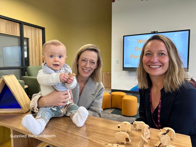 A mother and her baby smiling at the camera, as another woman sits beside them, also smiling. The baby is sitting on a timber shelf, and the walls behind them are yellow and white, with a TV mounted to the wall.