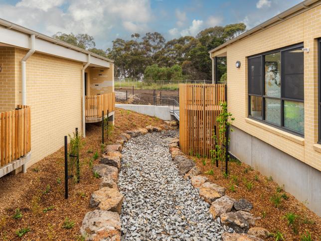 A man-made dry creek bed, made from gravel and larger rockets, running between two buuldings which have yellow brick walls and timber-fenced balcony areas