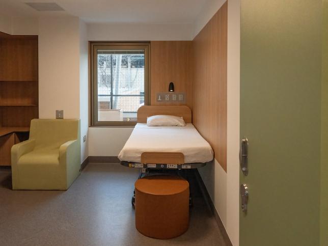 A room with timber and white walls, containing a mental health bed, stool, chair and shelf space.