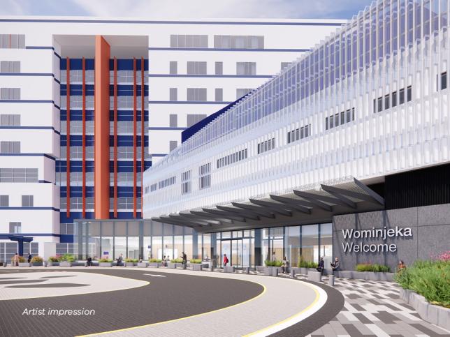 An artist impression of an external view of a hospital entrance. The entrance is made of glass, and a "Wominjeka, Welcome" sign is visible. The hospital building with white facade is visible in the background.e