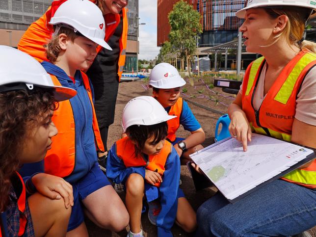 Four children in hi-vis vests and hard hats looking at a map. An adult is holding the map and pointing at it