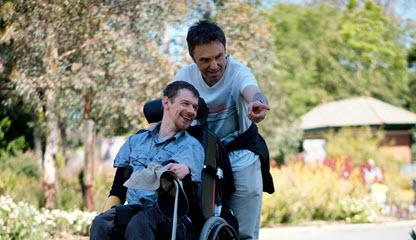 Image of an adult standing with a young person in a wheelchair, smiling