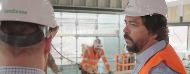 Trent Burke wearing a hard hat and hi vis construction vest at a worksite. He is standing with another man wearing a hard hat facing away from the camera.