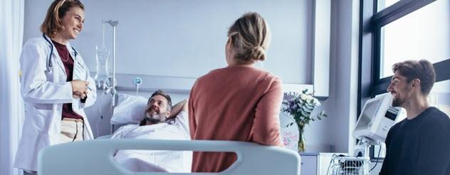 A doctor standing and talking to a patient in bed. There is a woman sitting on the patient's bed and a man sitting on a chair.