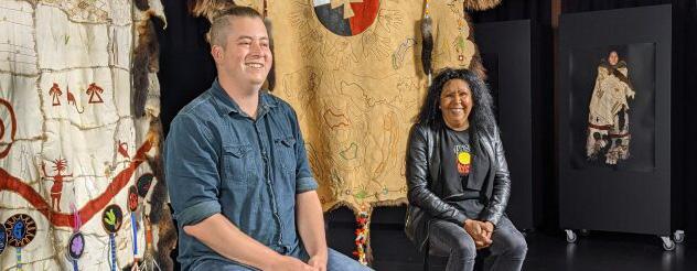 Tapestry artists Mitch Mahoney (left) and Maree Clark (right) sitting infront of their tapestries, smiling.