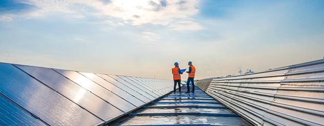 Two people wearing high visibility jackets stand on a roof, with a row of solar panels on their left