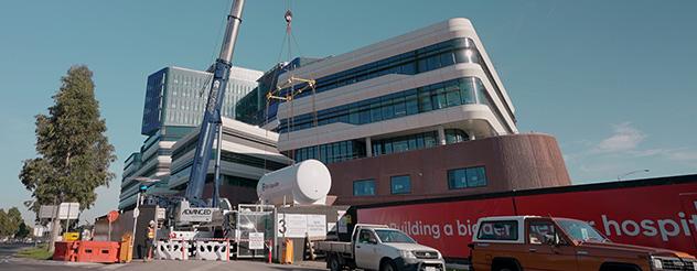 A crane lifting an oxygen tank, with the facade of the new Footscray Hospital visible behind it