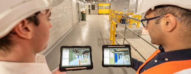 Two people wearing hard hats, holding tablets and looking at digital building models on the screens