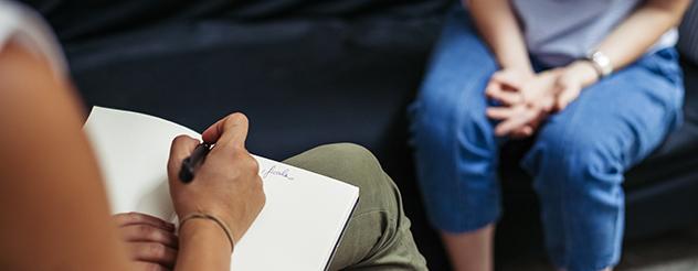 A person writing notes while facing another person sitting on a couch and speaking to them