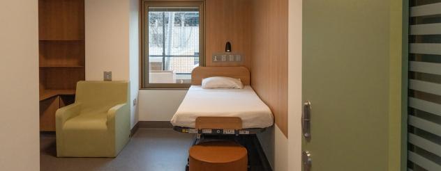 A room with timber and white walls, containing a mental health bed, stool, chair and shelf space.